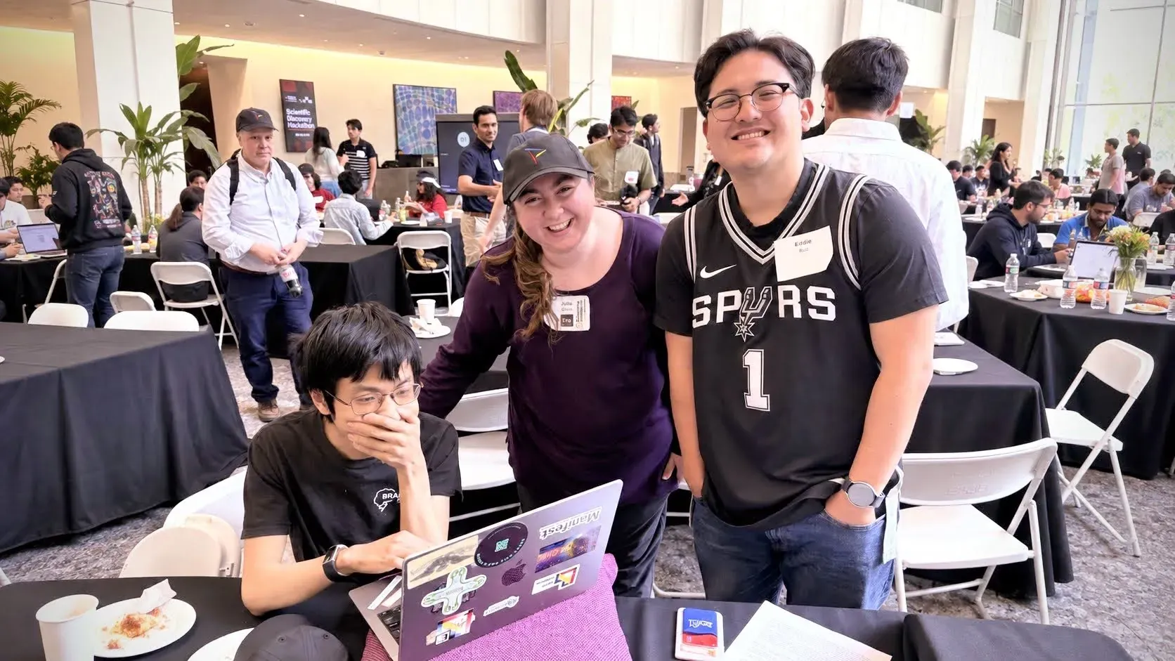 Julia Gross smiling with two fellow builders at a busy hackathon