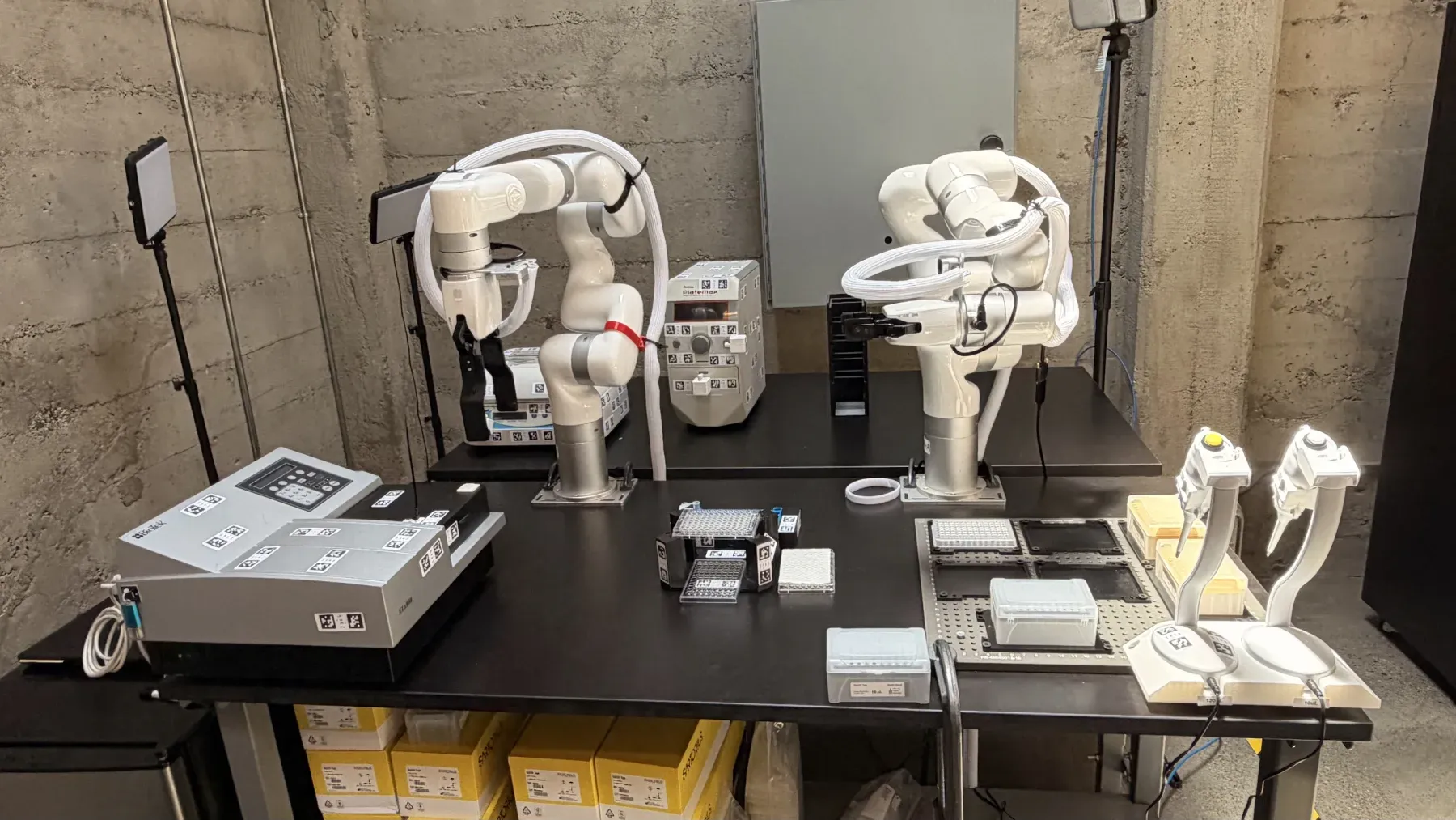 Two robotic arms at a lab bench with a plate reader, plate hotel, microplates, and pipettes staged around them
